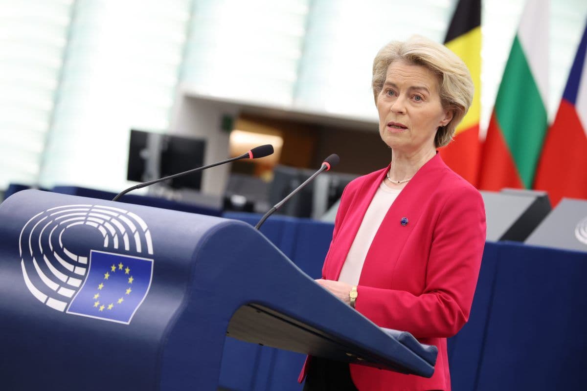 En person i en rosa blazer taler ved et podium med EU-merking. Flagg er synlige i bakgrunnen.