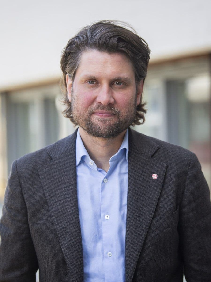 En mann med mellomlangt brunt hår og skjegg, iført blå skjorte og mørk blazer, står utendørs foran en bygning med vinduer.