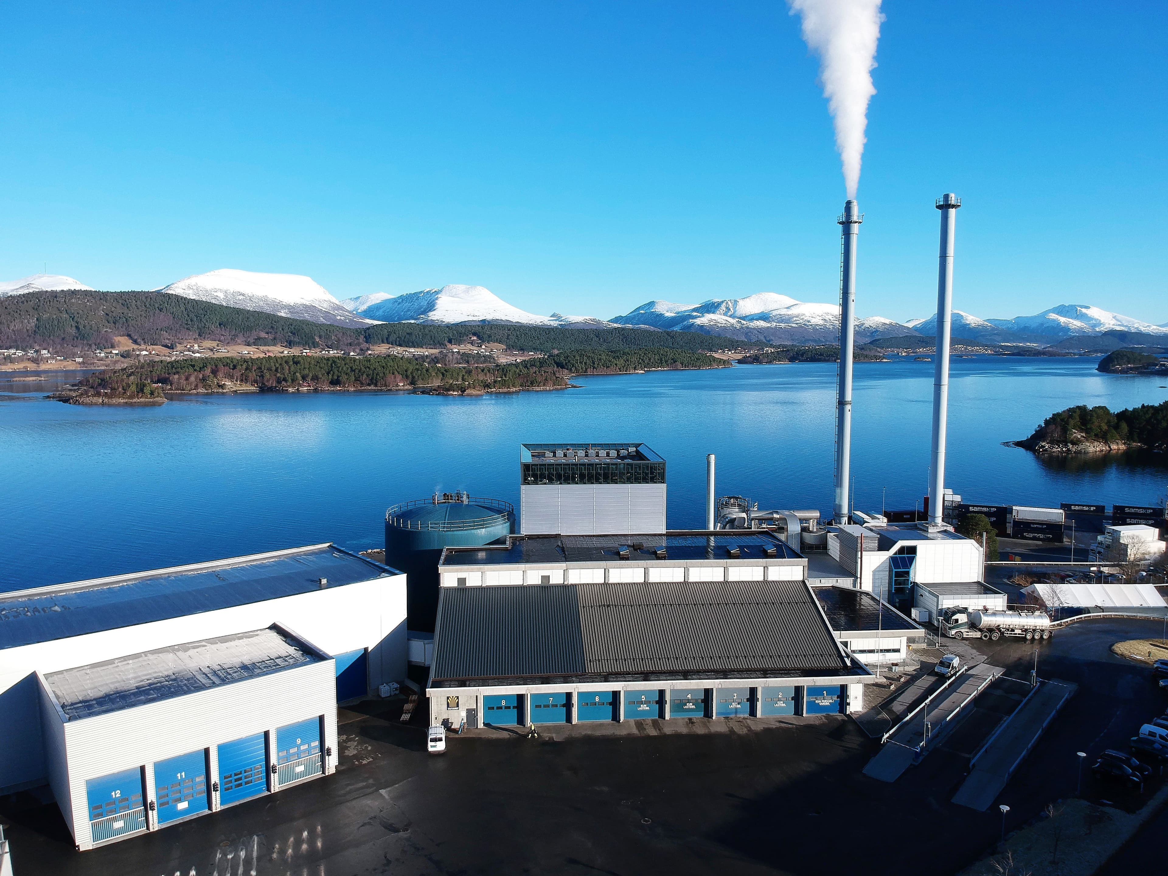 Industrianlegg ved vannkanten med to skorsteiner, hvorav den ene slipper ut røyk, med snødekte fjell i bakgrunnen.