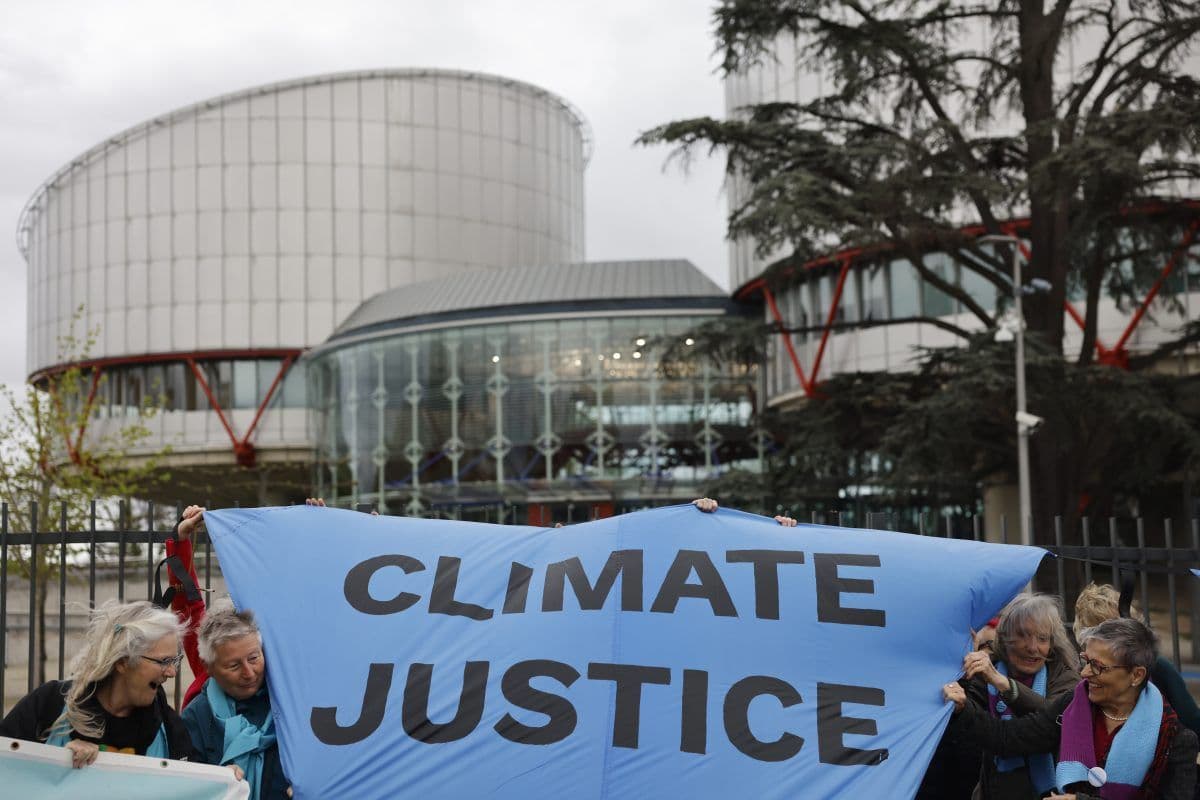 En gruppe mennesker holder et blått banner med teksten "CLIMATE JUSTICE" utenfor en stor, moderne bygning med sylinderformede strukturer.