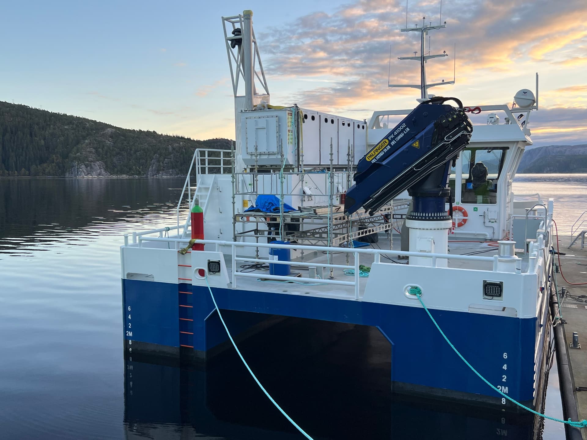 Blå og hvit katamaran-arbeidsbåt med kran og containere fortøyd i rolig vann, med skogkledde fjell i bakgrunnen.