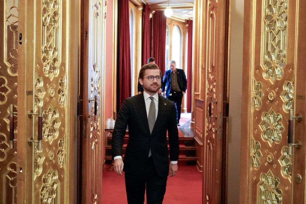 En mann i dress går gjennom utsmykkede gulldører i en elegant gang, med andre mennesker synlige i bakgrunnen.