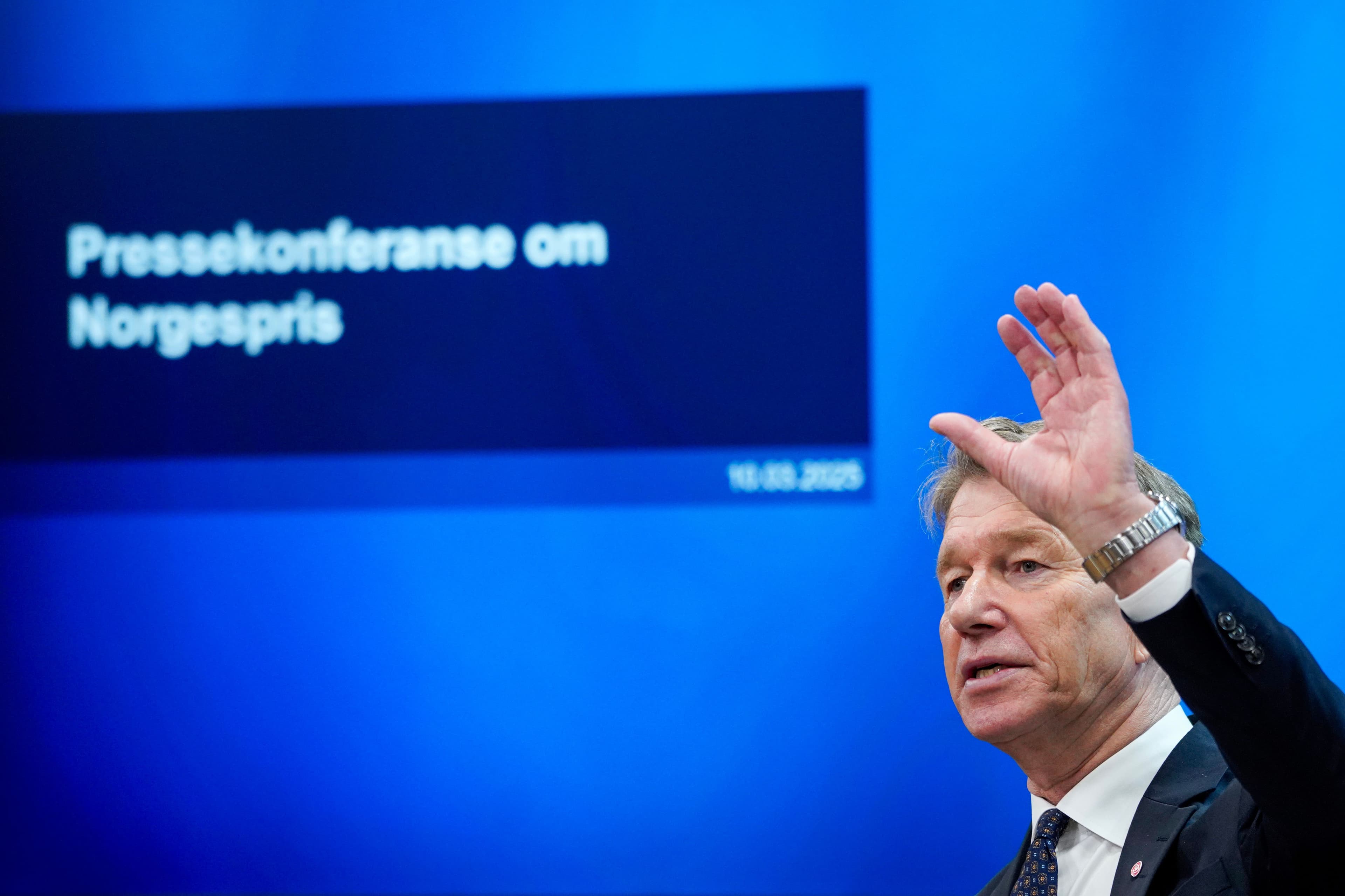 En mann med hånden hevet snakker foran en blå skjerm med teksten «Pressekonferanse om Norgespris».