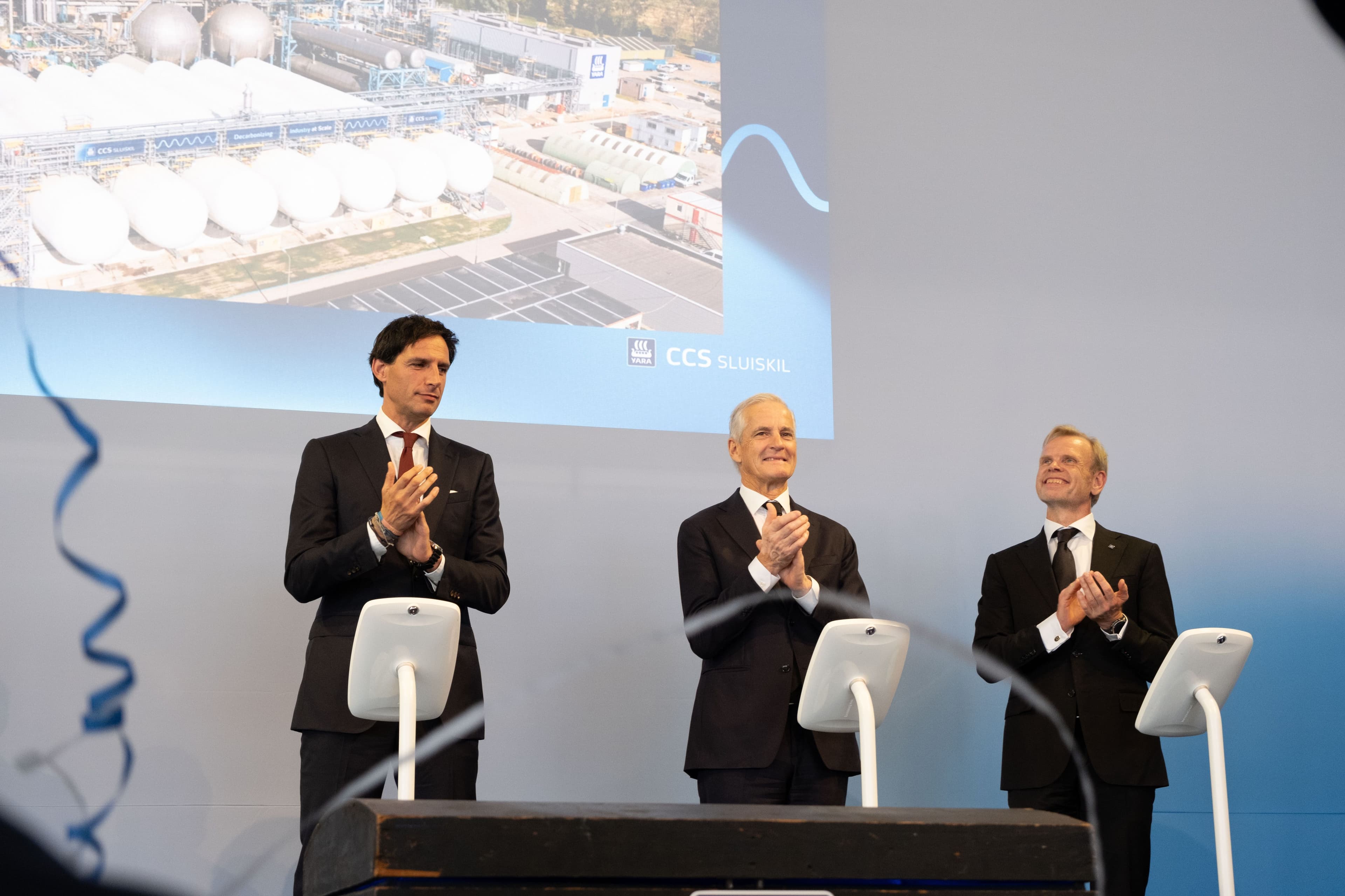 Tre menn i dress klapper på en scene foran en skjerm som viser et industrianlegg og teksten «CCS SLUISKIL».