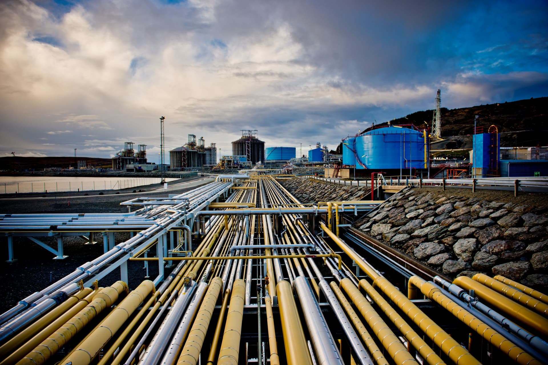 Rørledninger i forgrunnen fører til et industrianlegg med store blå lagertanker og andre bygninger under en overskyet himmel.