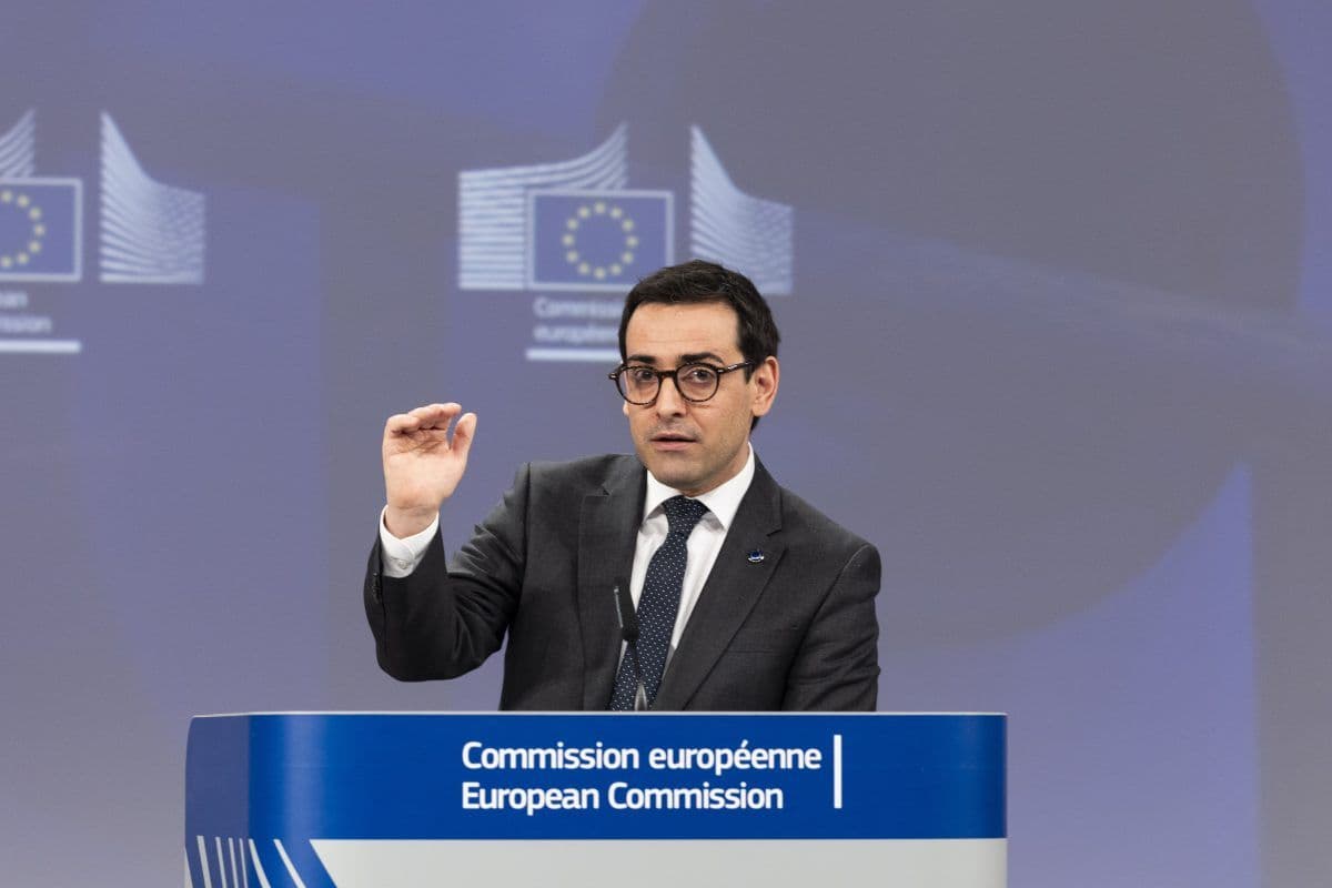 En person i dress snakker ved et podium med EU-kommisjonens logoer og gestikulerer med én hånd.