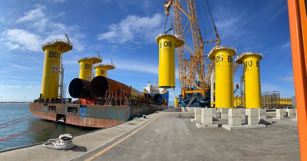 Store, gule, sylinderformede konstruksjoner merket B01 til B08 løftes med kraner fra kaikanten og over på et skip i et industrielt havneanlegg.