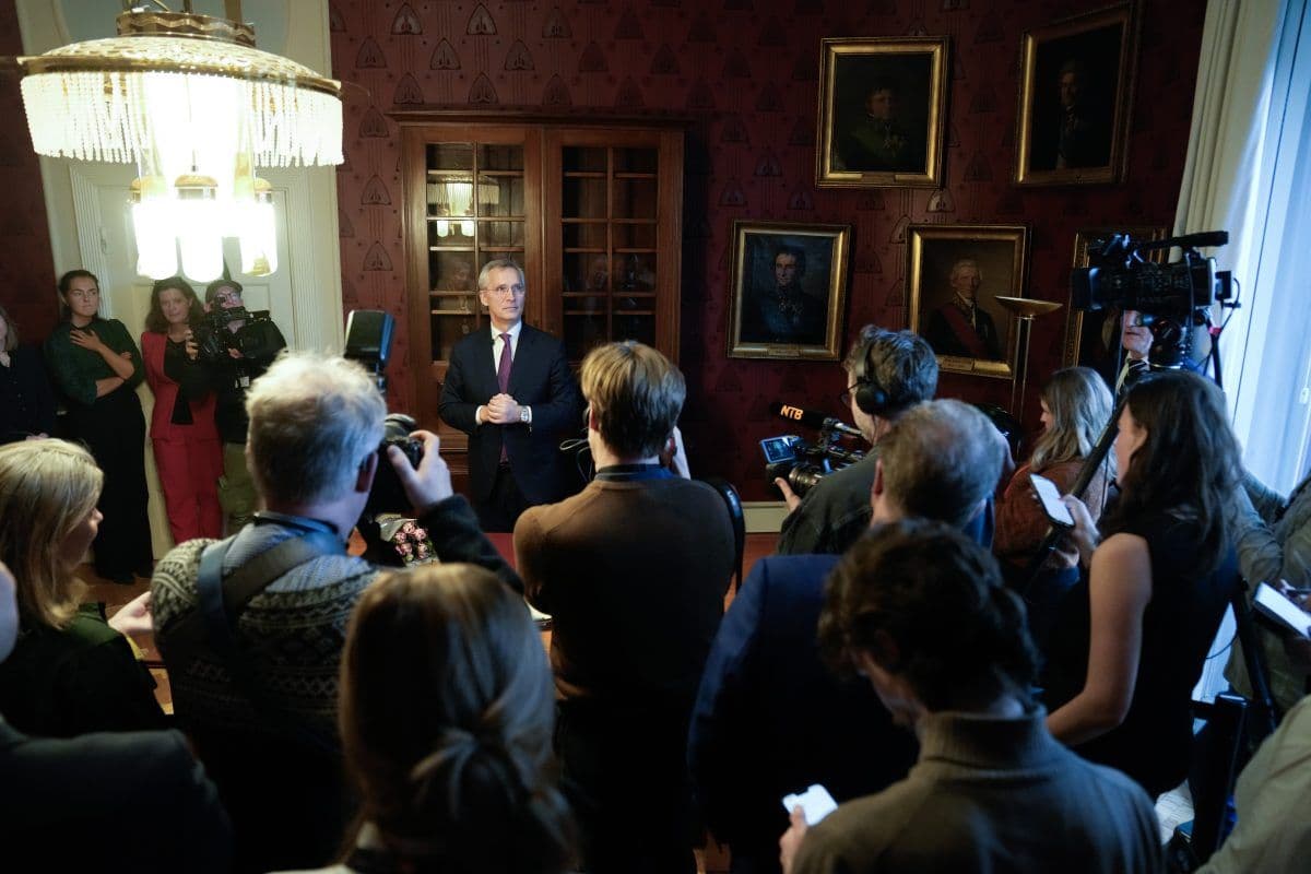 En gruppe mennesker og fotografer omgir en mann som snakker i et rom med innrammede portretter og en stor lysekrone.