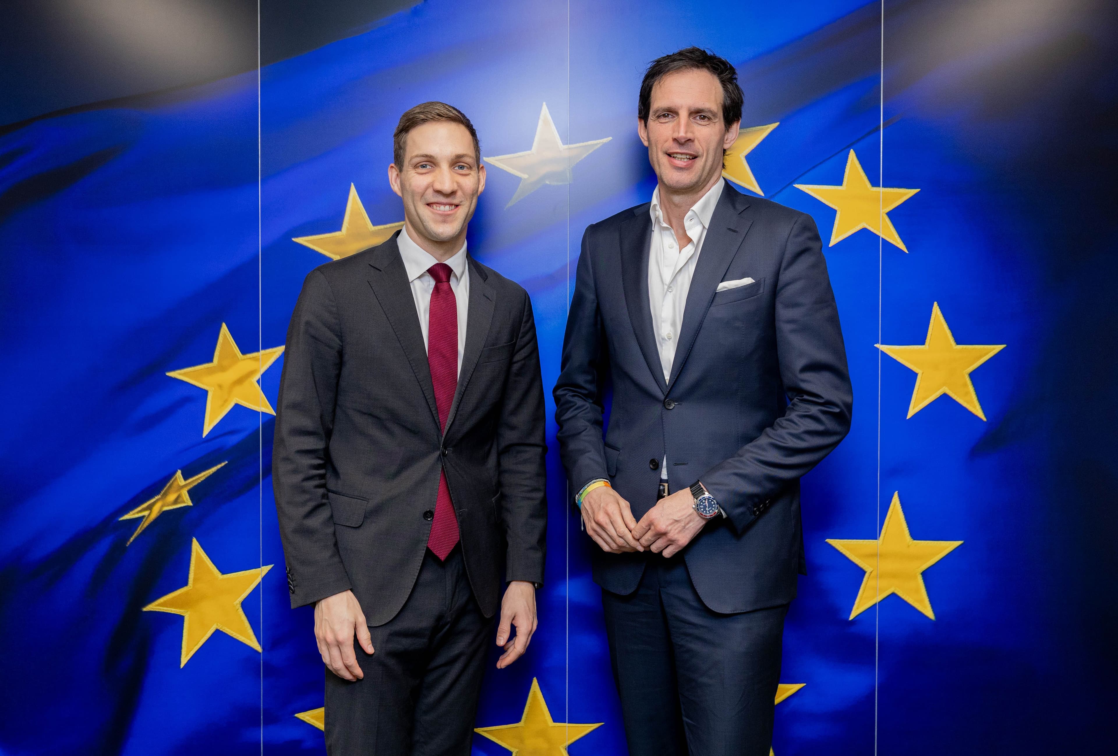 Two smiling men in suits stand in front of a European Union flag.