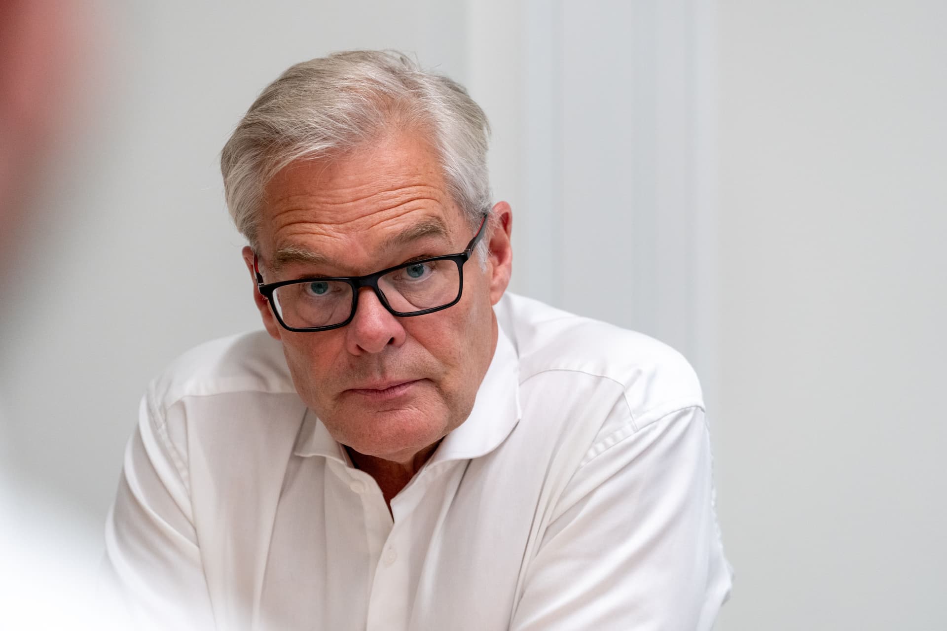 En mann med grått hår og briller i hvit skjorte ser rett inn i kameraet.