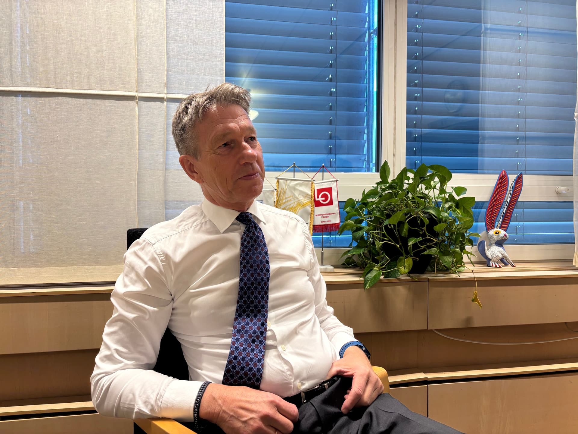 En mann i hvit skjorte og blått slips sitter og ser mot høyre, med et vindu, en plante og en fuglefigur bak seg.