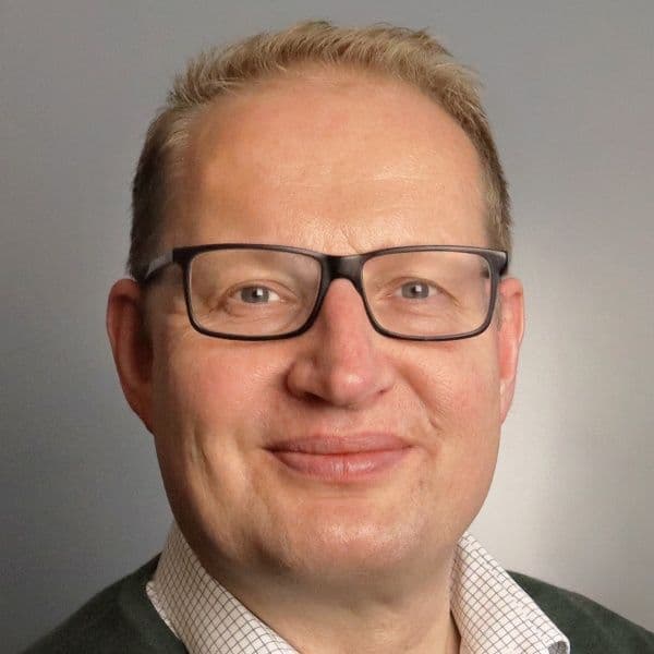 En person med briller, kort blondt hår og iført skjorte med krage smiler mot en vanlig grå bakgrunn.