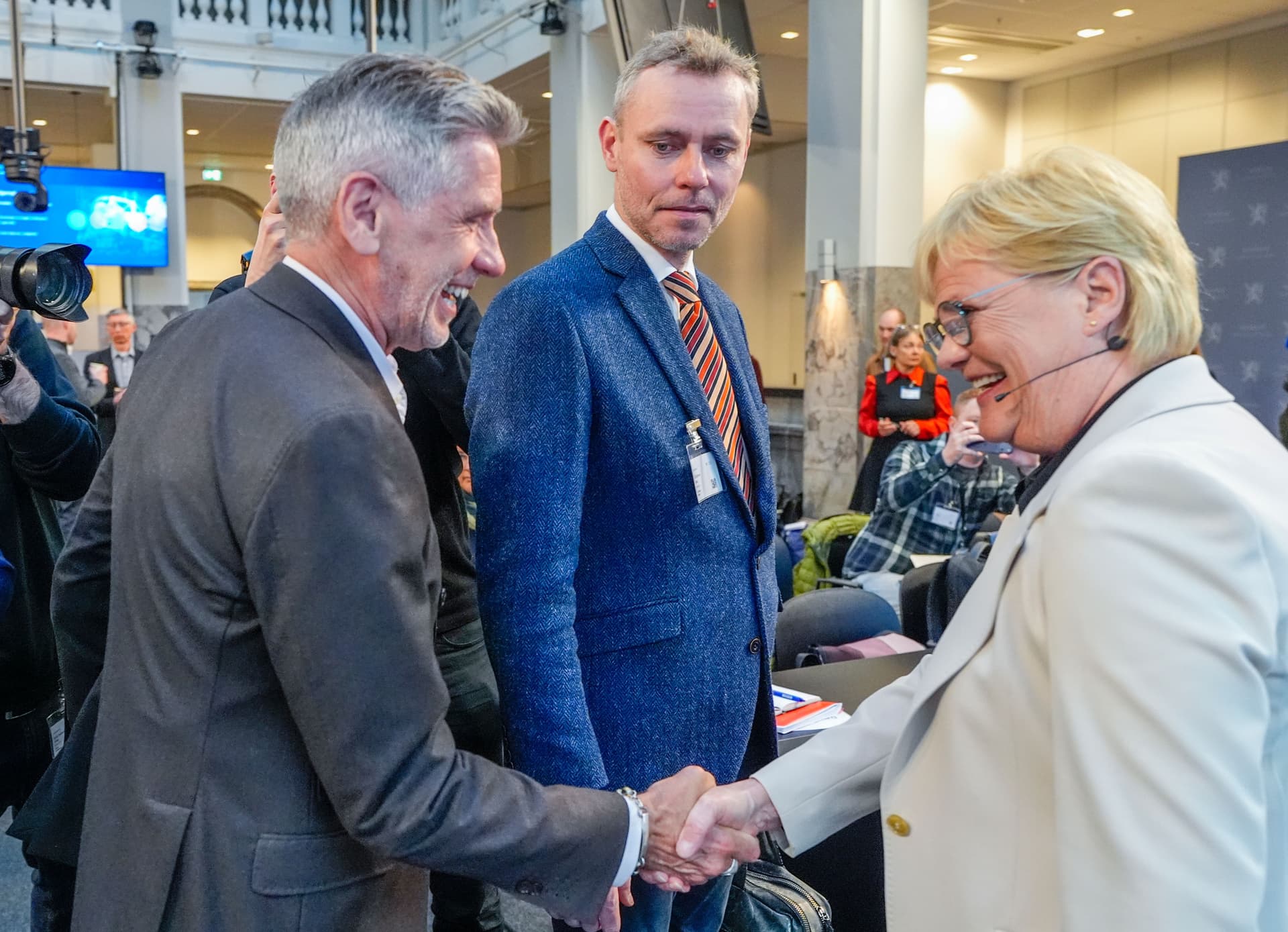 En mann og en kvinne smiler mens de håndhilser, med en annen mann som observerer, på et innendørs arrangement.