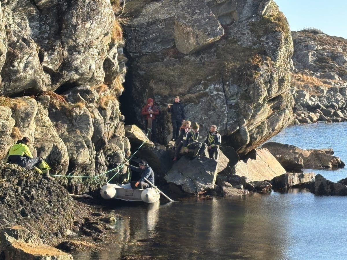 En gruppe mennesker i friluftsutstyr sitter og står på steiner ved en strandlinje, med en liten gummibåt knyttet til et tau i nærheten.