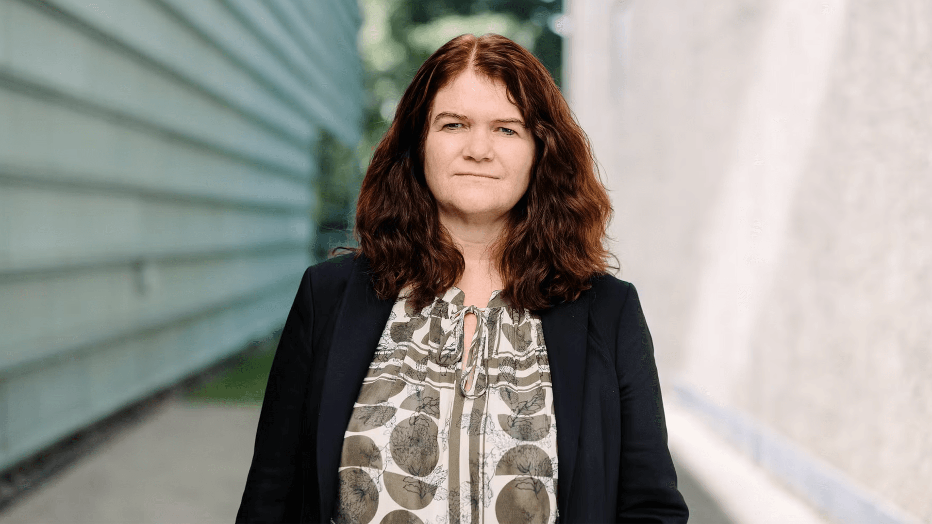 En kvinne med rødbrunt hår, iført en svart blazer og en mønstret bluse, står utendørs.