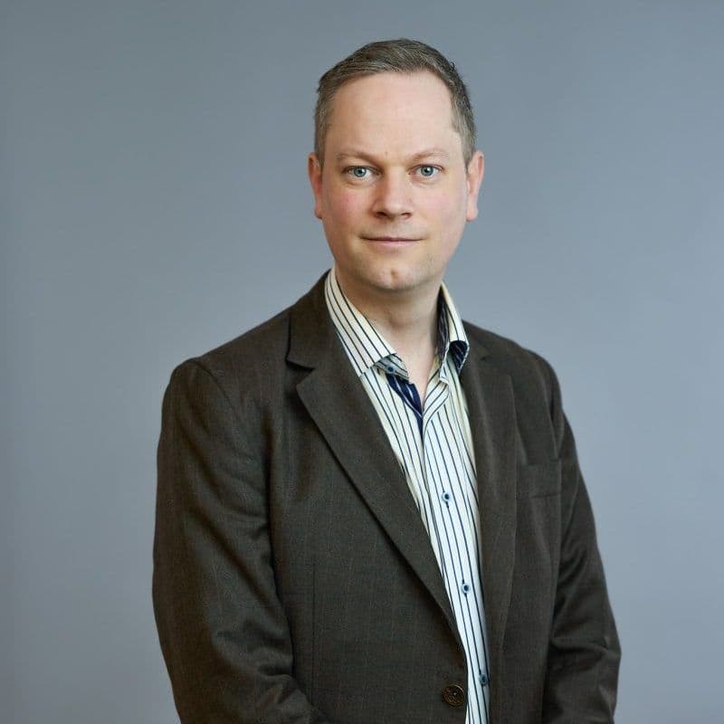 Person i brun blazer og stripet skjorte som står mot en vanlig grå bakgrunn.