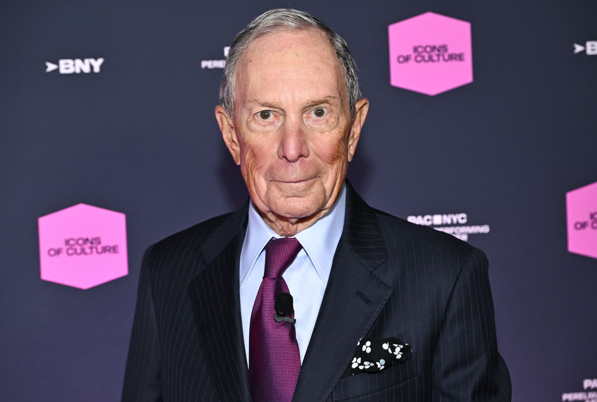 Michael Bloomberg i mørk dress og lilla slips, stående foran en mørk bakgrunn med rosa «Icons of Culture»-logoer.