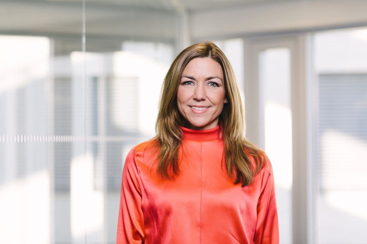 En kvinne med langt lysebrunt hår iført en skinnende rød bluse står innendørs foran store vinduer og glassvegger og smiler til kameraet.