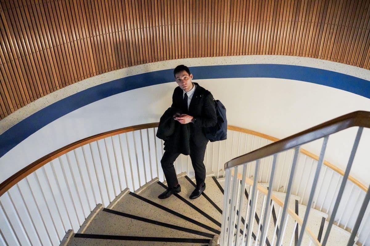 Mann i formell antrekk står på en spiraltrapp, holder en frakk og ser på telefonen sin. Trappen har hvitt rekkverk og en blå stripe på veggen.