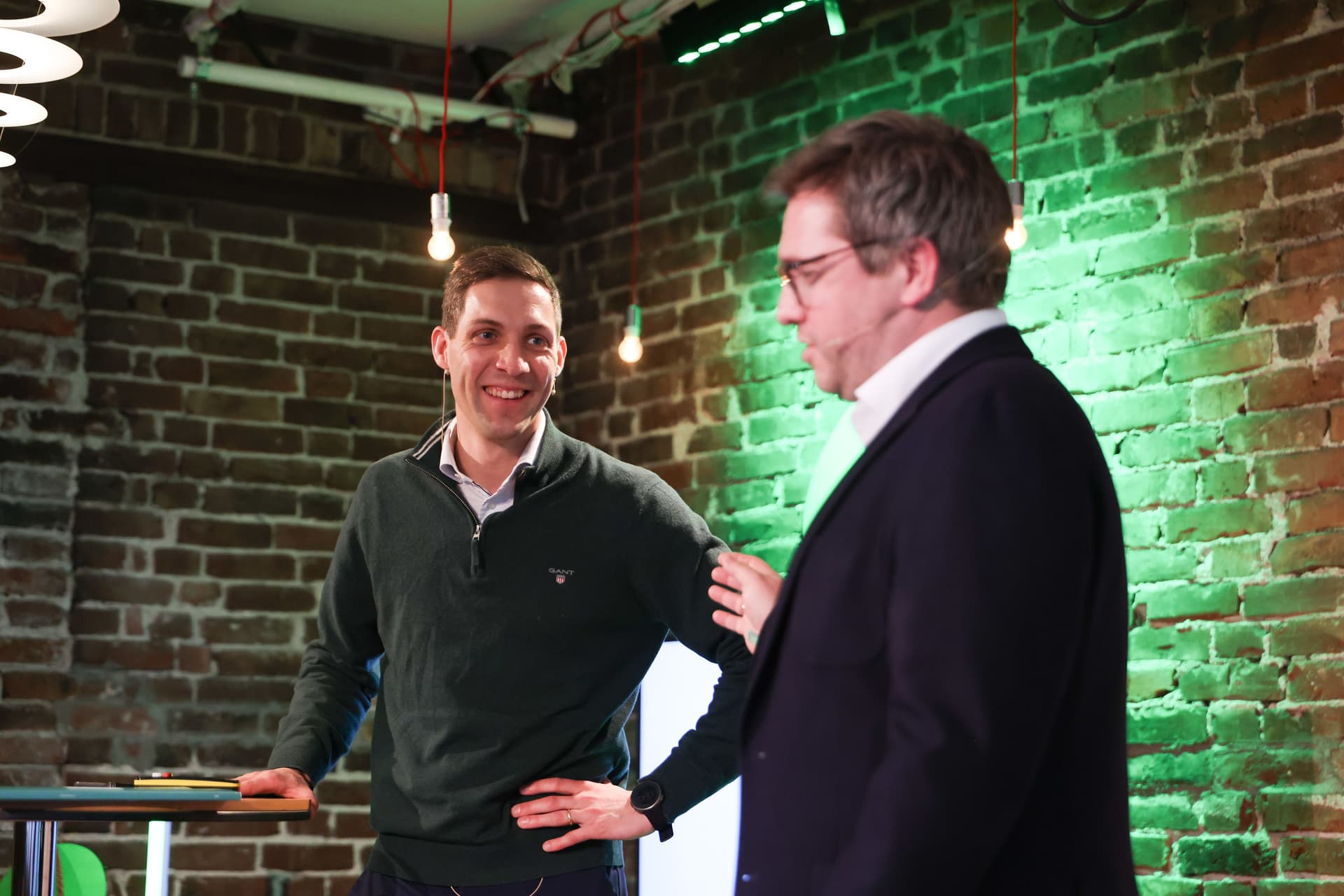 Man in a green sweater smiles at a man in a suit, with a brick wall background.