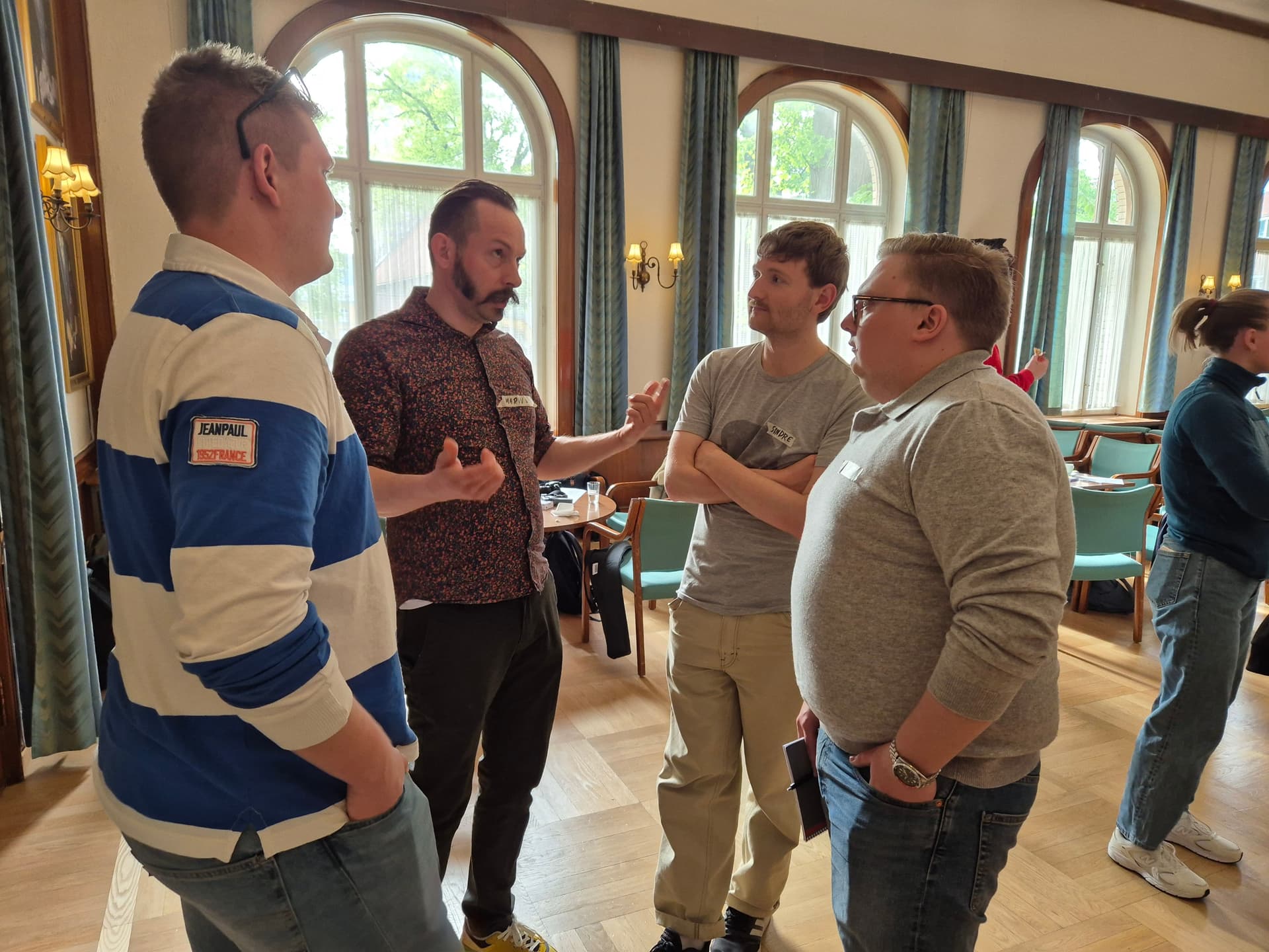 Fire menn i diskusjon i et rom med buede vinduer.