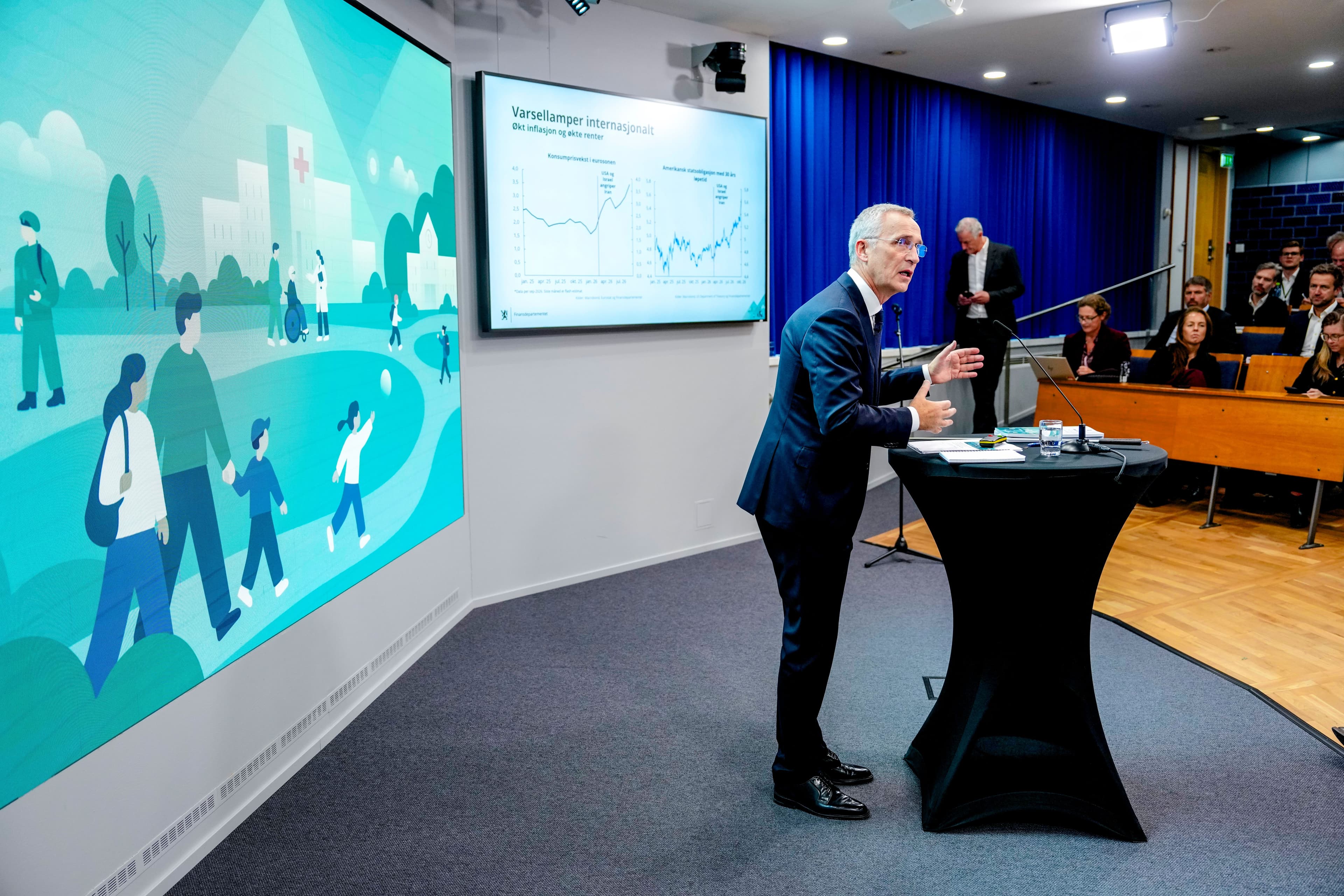 A man in a suit presents at a podium to an audience, with screens behind him showing a graph and an illustration of people and a hospital.