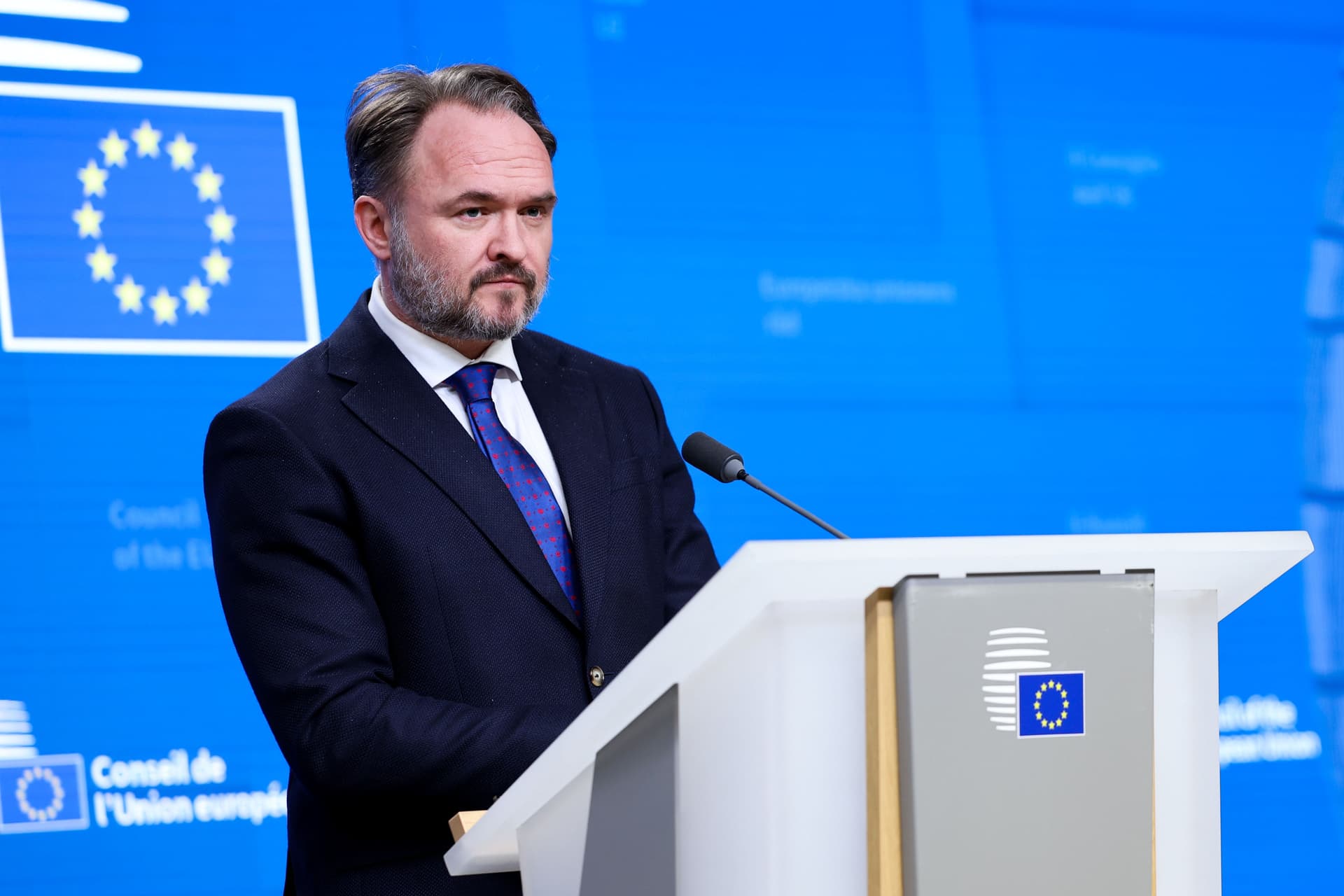 En skjeggete mann i mørk dress taler fra en talerstol med EU-logo, foran en blå bakgrunn med EUs flagg.