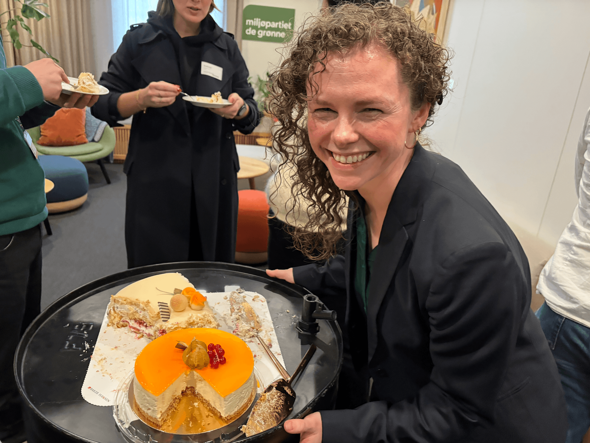 En smilende kvinne holder et brett med delvis oppspiste kaker, mens andre mennesker spiser i bakgrunnen og et skilt med teksten «Green Party» vises.