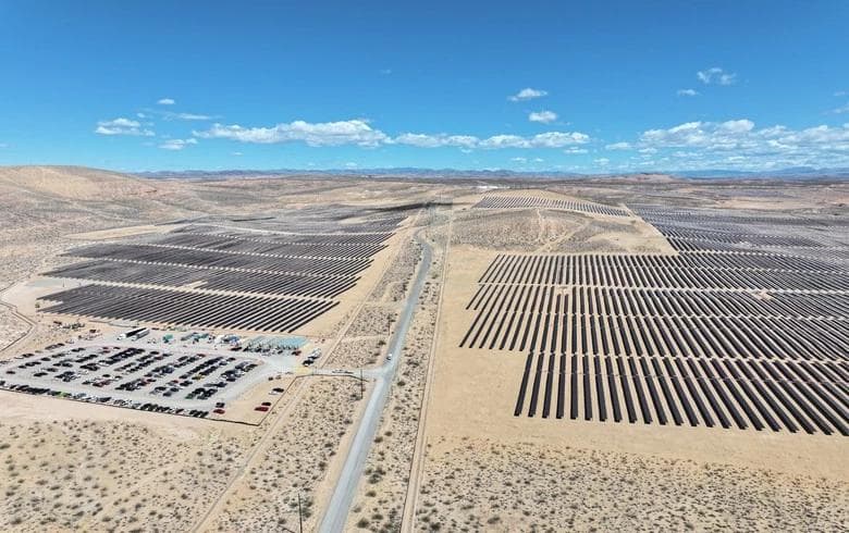 Flyfoto av en stor solcellepark i et ørkenlandskap, med rader av solcellepaneler og en parkeringsplass med mange biler.