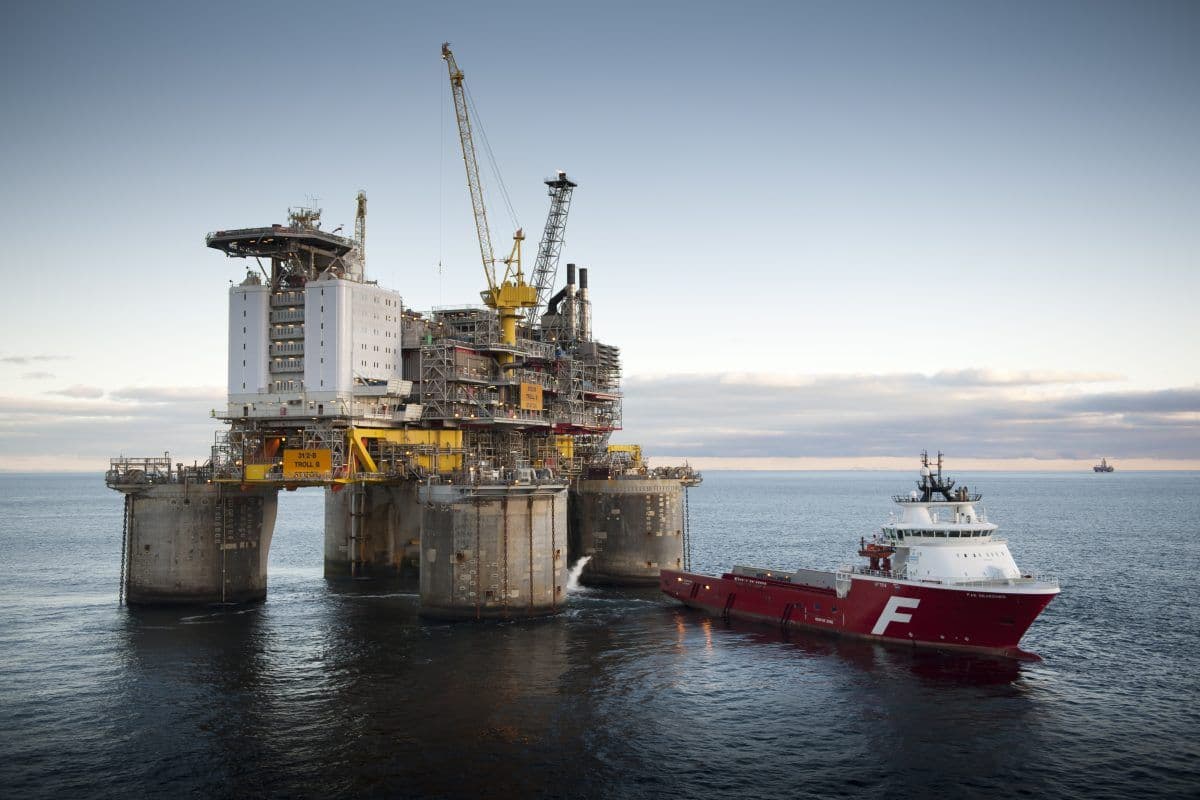 En oljeplattform til havs står i havet med et rødt forsyningsskip fortøyd ved siden av under en delvis overskyet himmel.