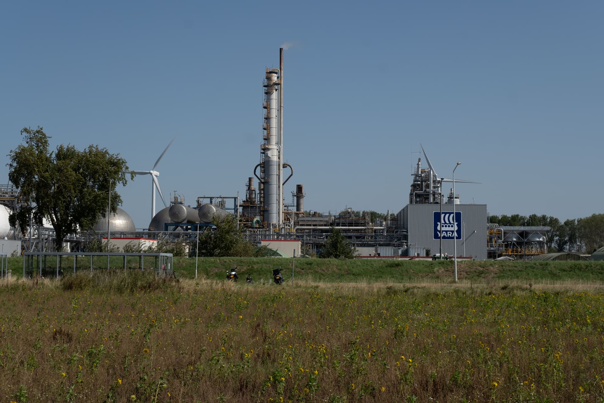 Yara industrial plant with towers, tanks, and wind turbines, beyond a field of dry grass.