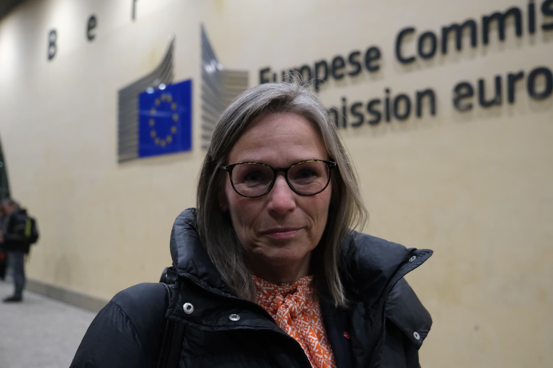En kvinne med grått hår og briller, iført en svart jakke, står foran logoen og teksten til Europakommisjonen.