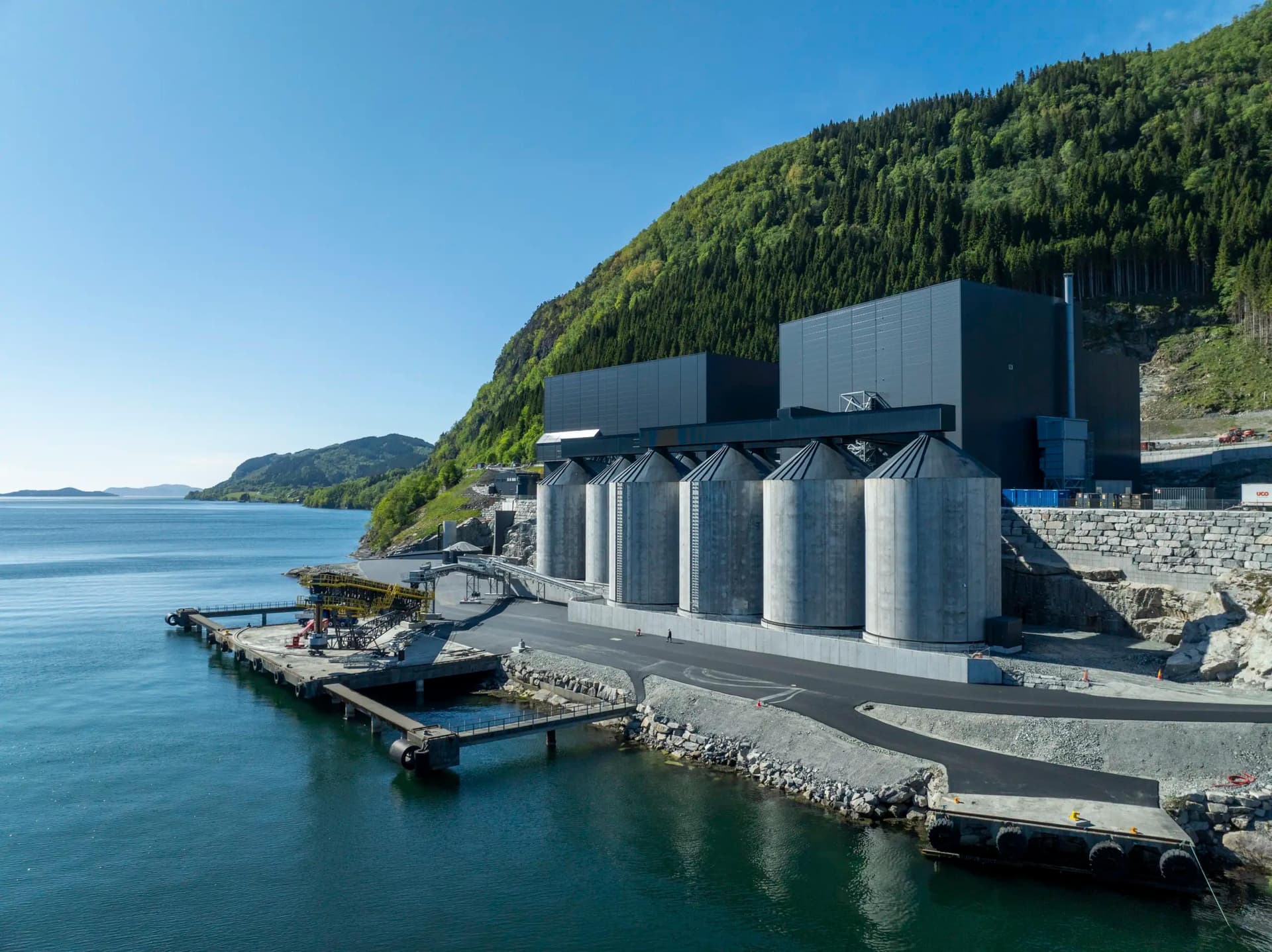 Moderne industrianlegg med store siloer og mørke bygninger på en kai ved en bukt, med en skogkledd fjellside i bakgrunnen.