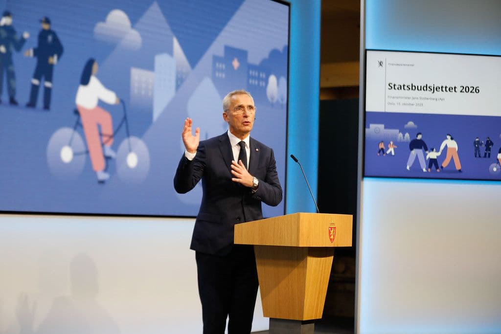 Jens Stoltenberg som taler ved en talerstol foran en skjerm med teksten «Statsbudsjettet 2026» og illustrasjoner av mennesker.