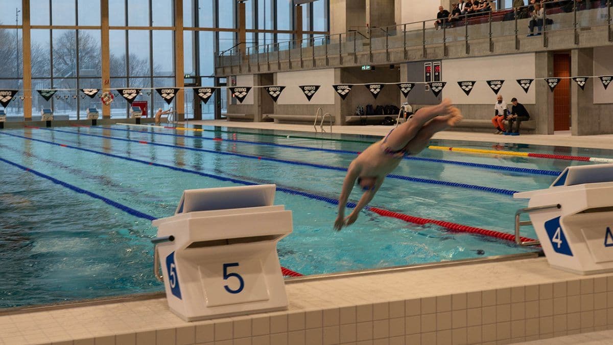 Svømmer stuper ned i et innendørsbasseng fra startblokken merket nummer 5, med synlige baner og noen få tilskuere i sittegruppen.