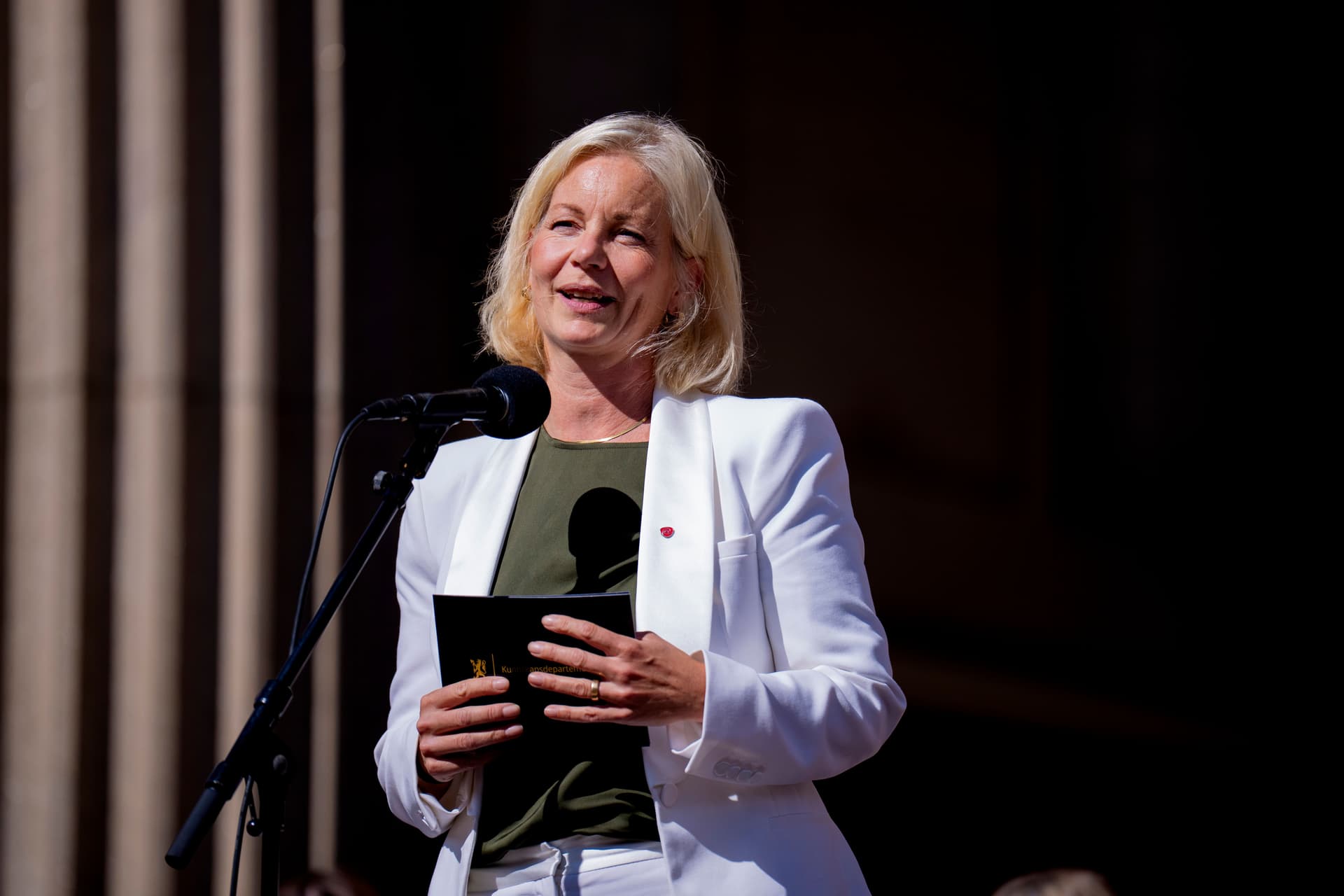 A blonde woman in a white blazer speaks into a microphone while holding a dark book.