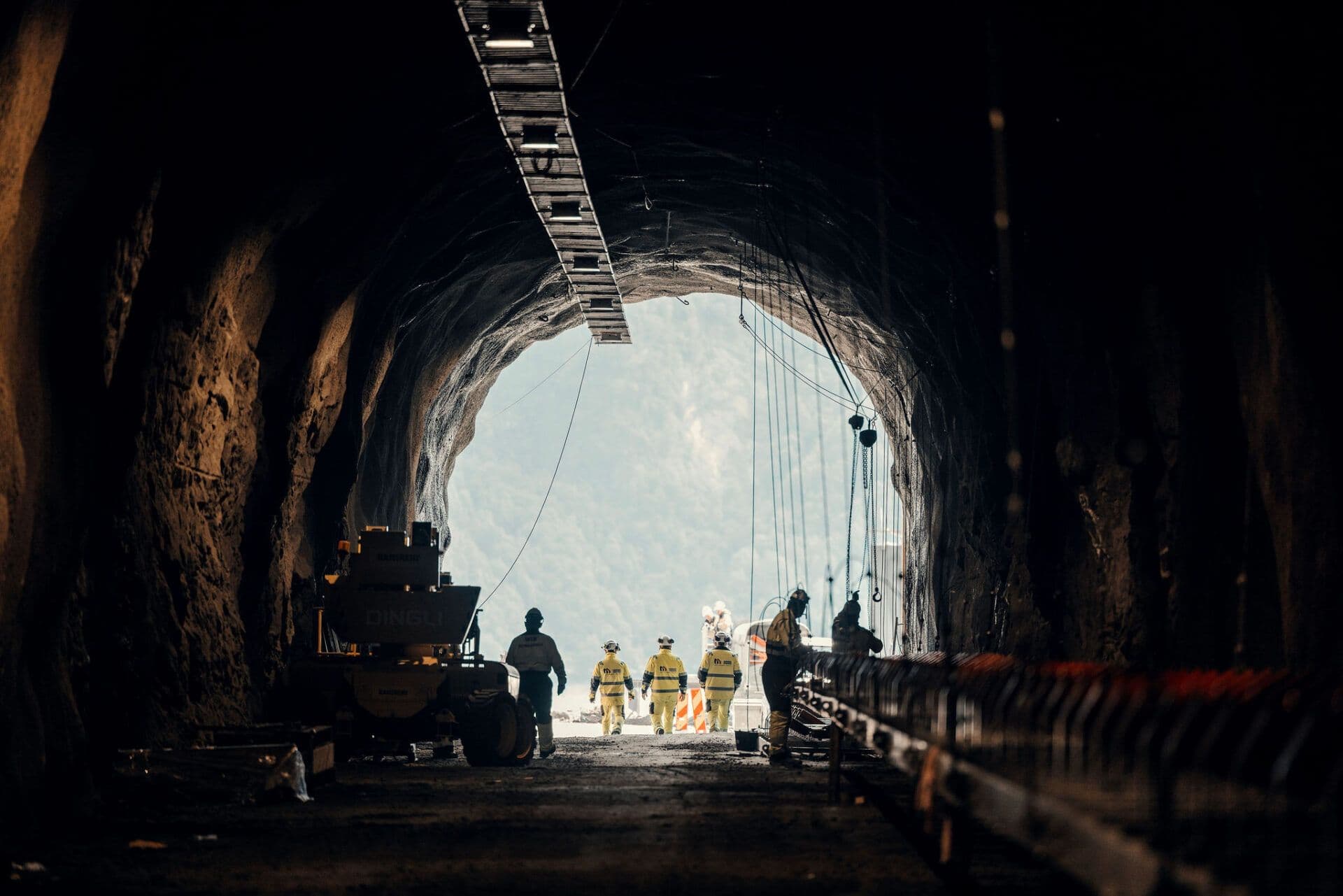 Bygningsarbeidere og utstyr inne i en mørk tunnel, vendt mot en lys utgang mot en fjelldal.