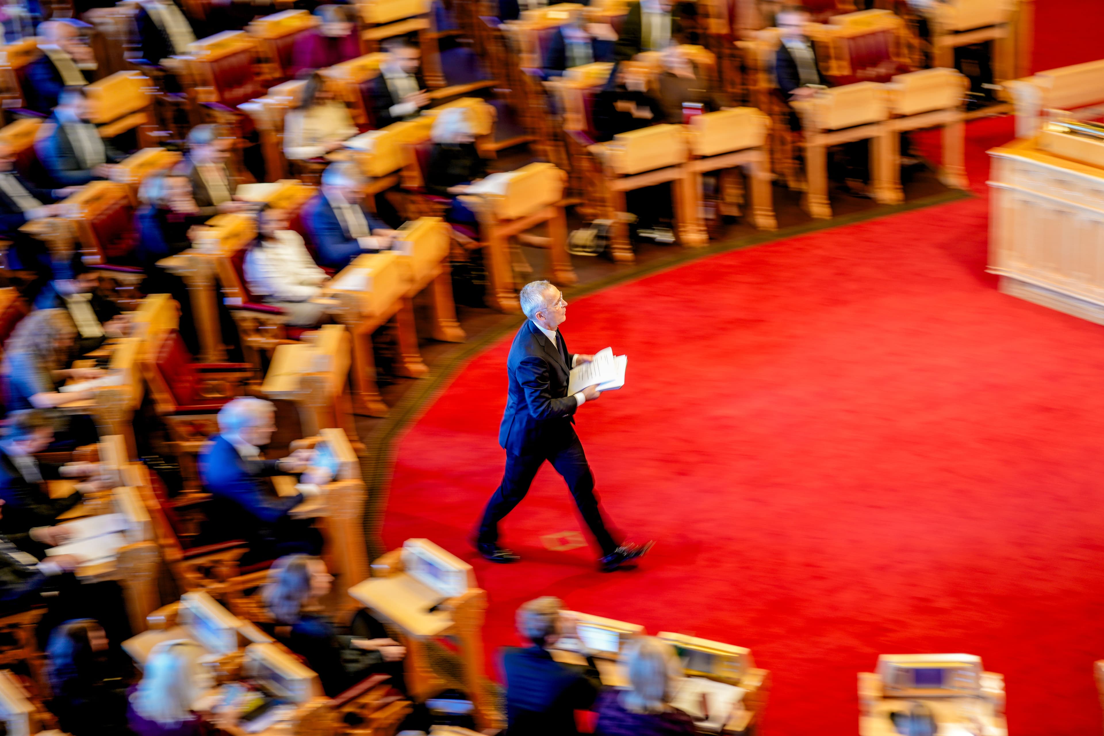 En mann i mørk dress går gjennom stortingssalen mens han holder noen papirer, med sittende deltakere ute av fokus i bakgrunnen.