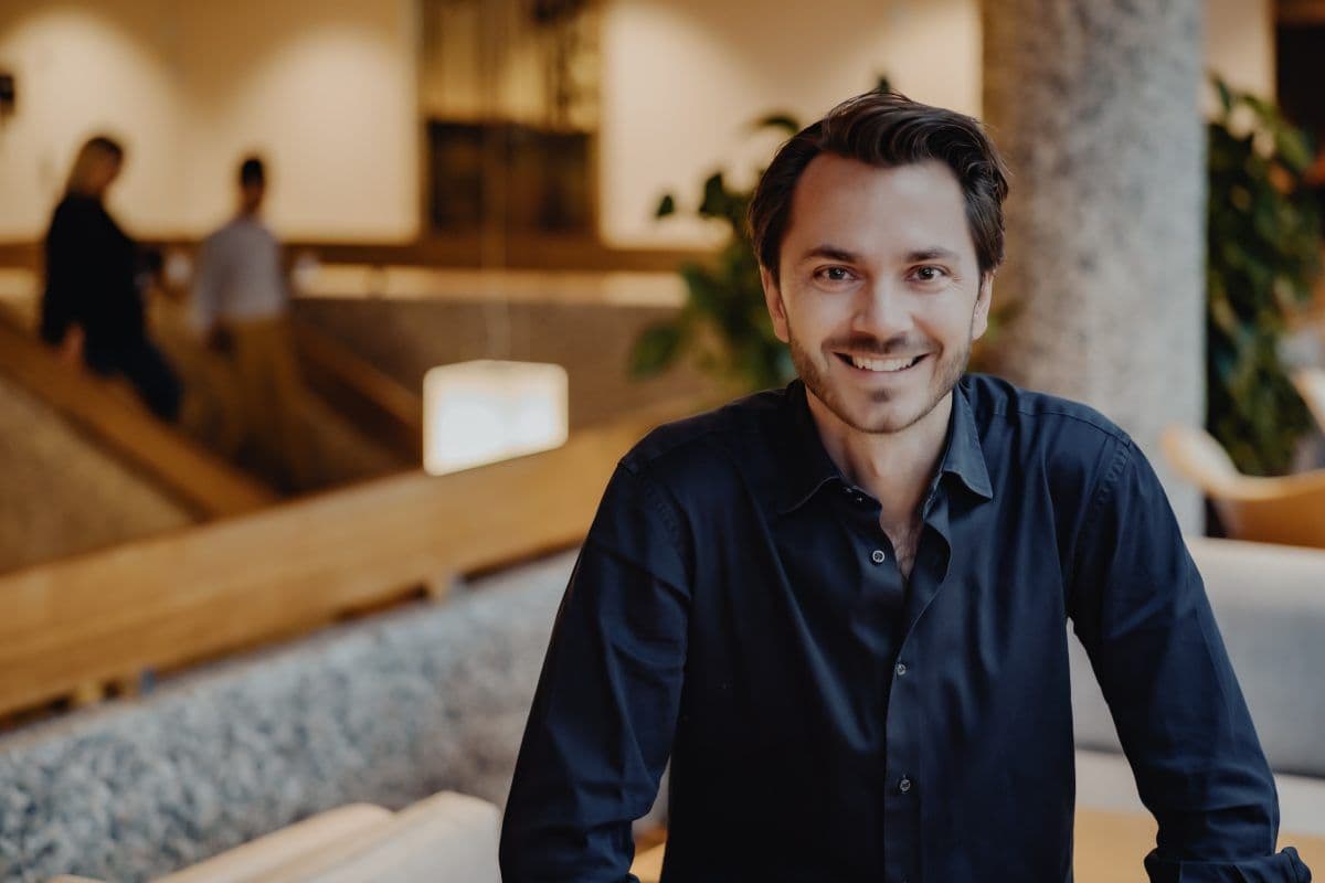 En mann med mørkt hår og mørk skjorte smiler mens han sitter innendørs; en uskarp bakgrunn viser to personer og moderne innredning.