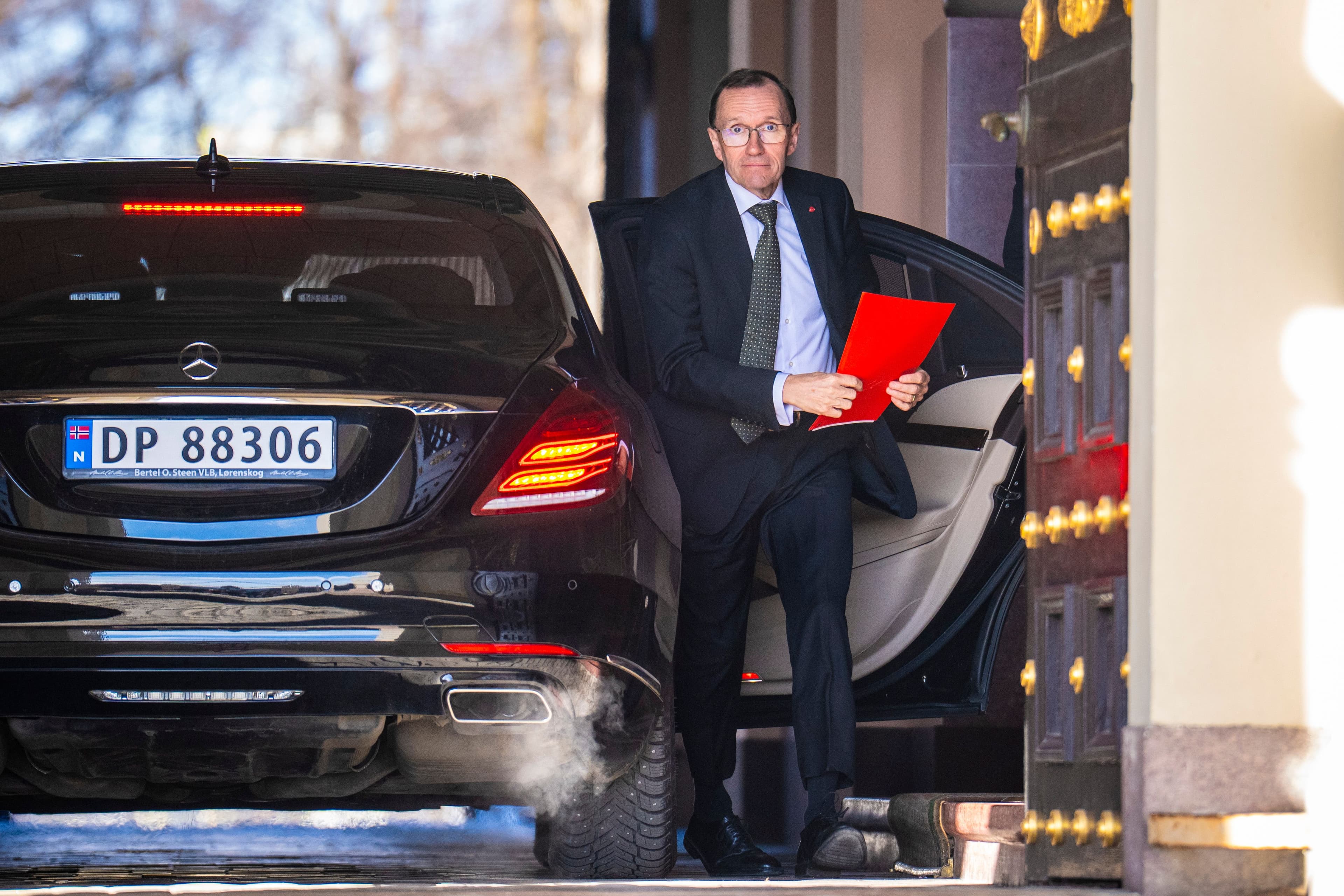En mann i dress stiger ut av en svart Mercedes-sedan og holder røde papirer.