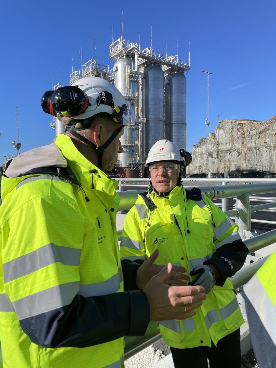 To arbeidere i synlige jakker og hjelmer diskuterer på en industriplass med store metalliske strukturer i bakgrunnen.