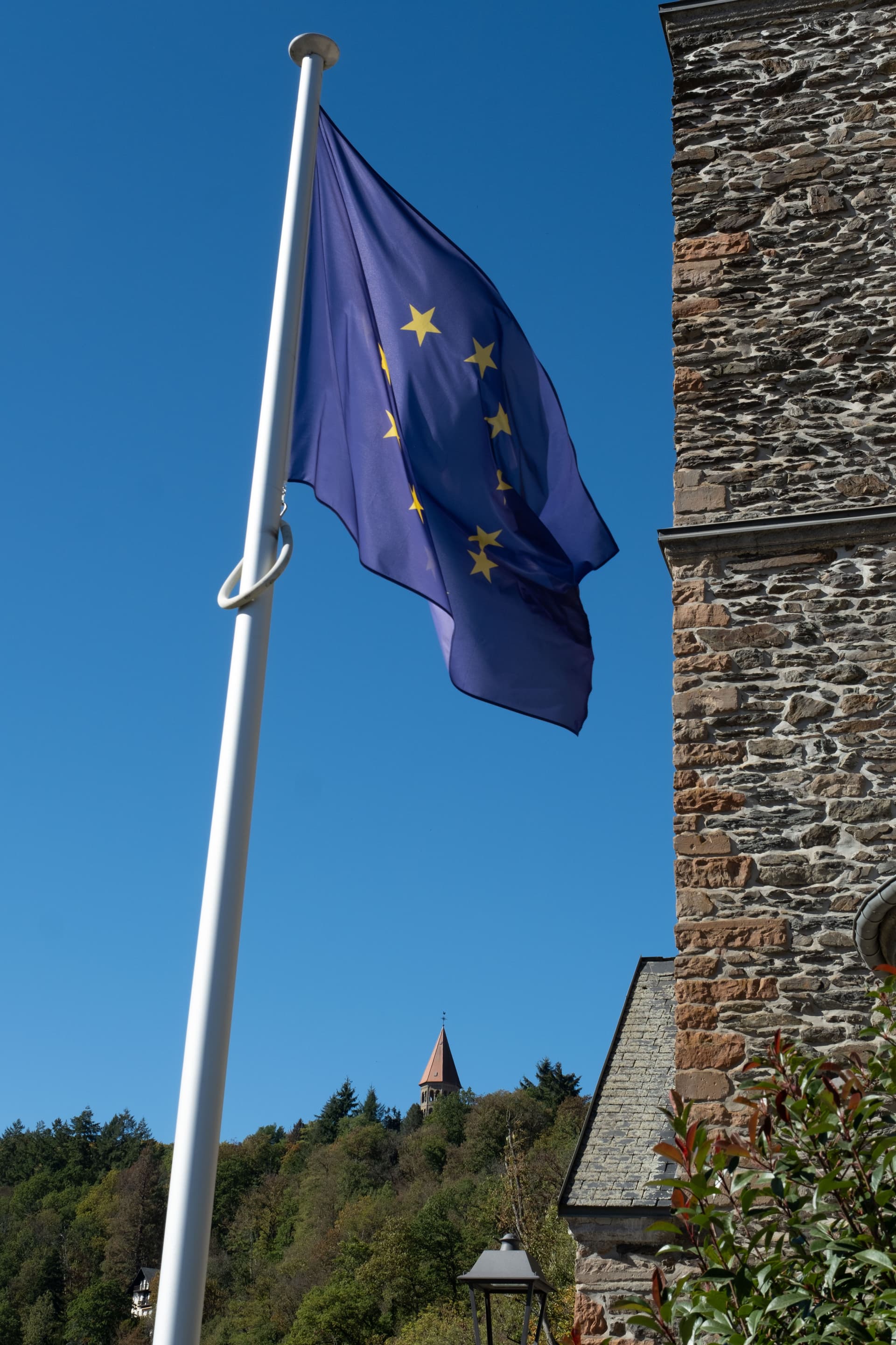 EU-flagget vaier mot en blå himmel ved siden av en steinbygning, med et tårn på en høyde i det fjerne.