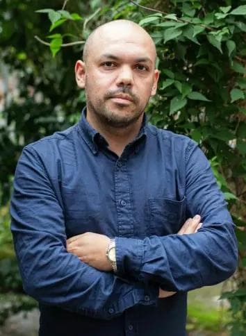 En mann med barbert hode og skjegg, iført en mørkeblå skjorte og klokke, med armene i kors foran grønn vegetasjon.