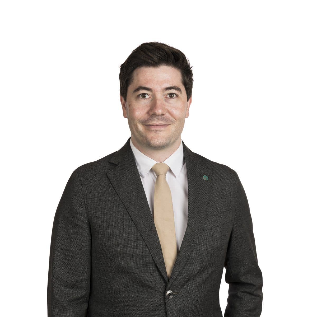 En mann i mørk dress, hvit skjorte og beige slips står vendt fremover mot en enkel, hvit bakgrunn.