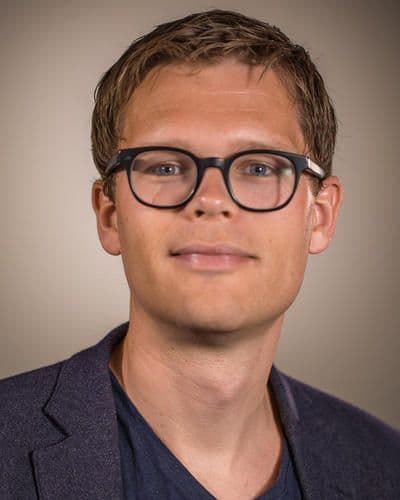 En mann med kort, lysebrunt hår, iført briller med svart innfatning, mørk blazer og blå skjorte, poserer foran en nøytral bakgrunn.