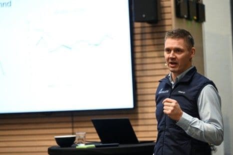 A man in a blue vest speaks, gesturing, in front of a large screen displaying a line graph.