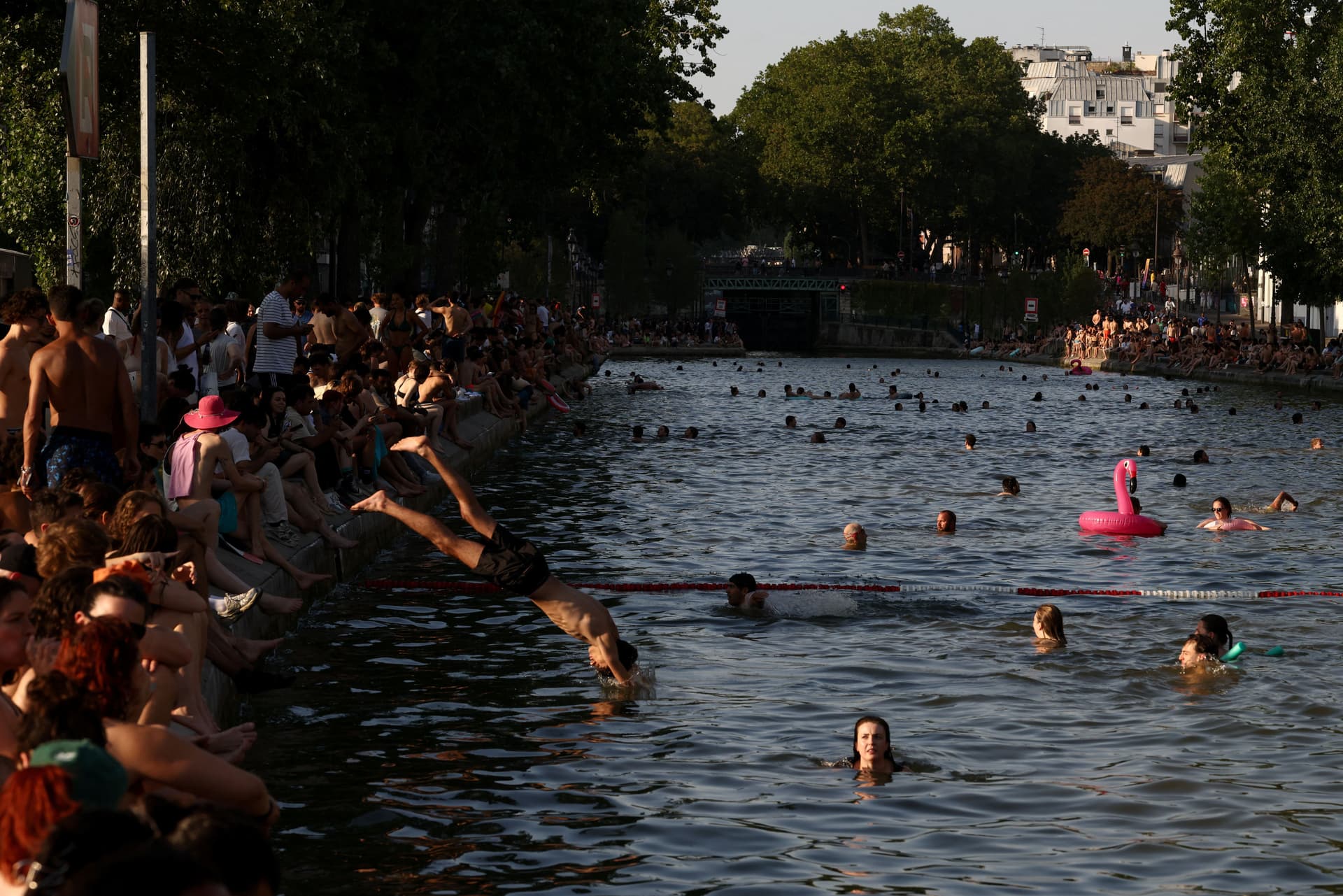 En travel kanalscene med folk som svømmer, dykker og slapper av på breddene, med en rosa flamingo-badeflåte i vannet.