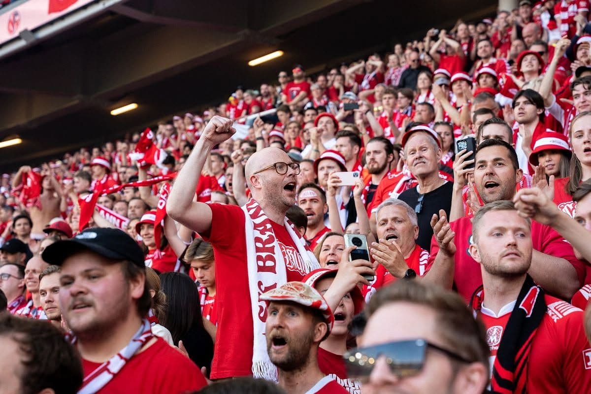 En stor folkemengde med entusiastiske supportere i røde trøyer og skjerf, med en mann i forgrunnen som løfter knyttneven og jubler.