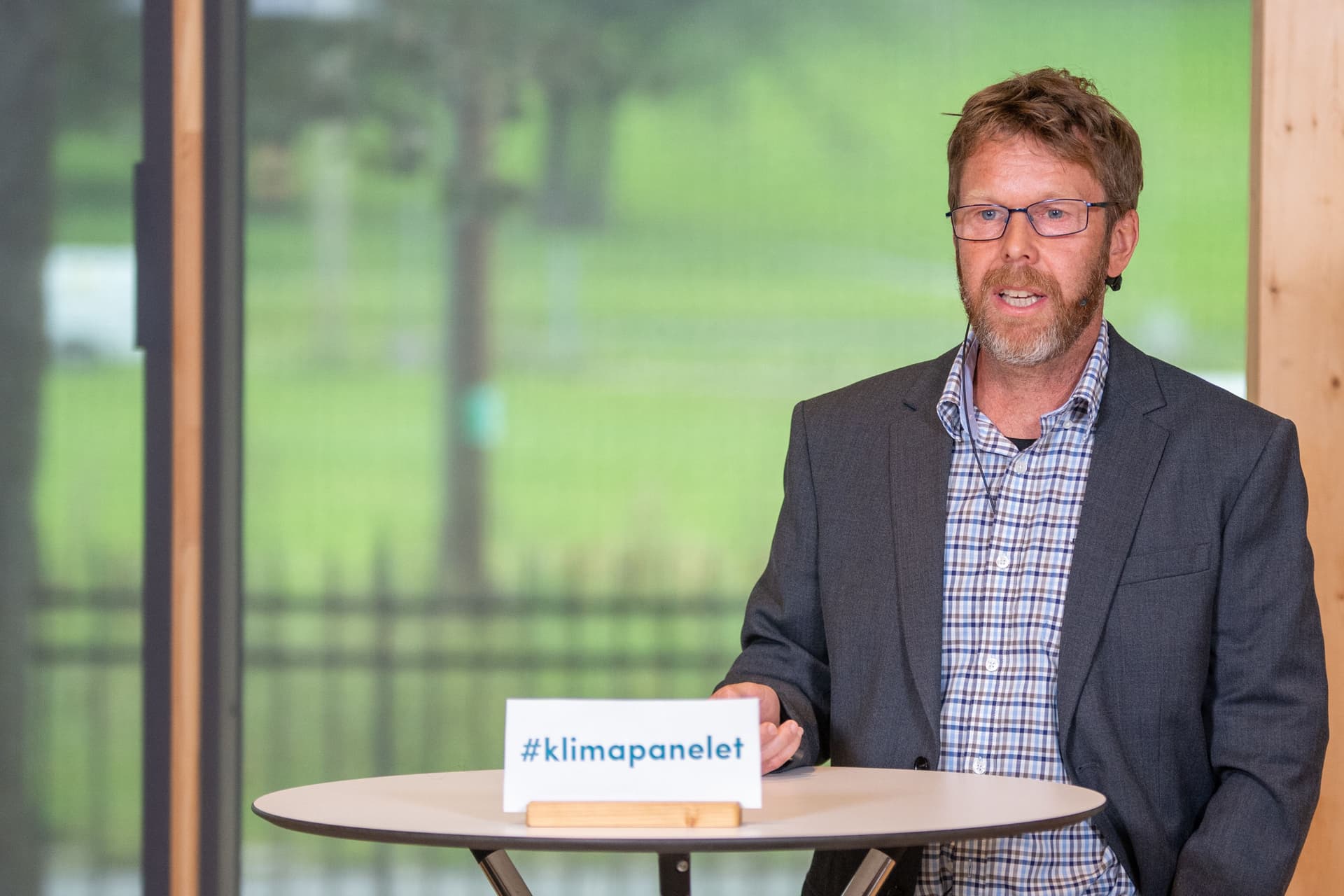 En mann med briller og skjegg snakker bak et bord med et skilt hvor det står «#klimapanelet».