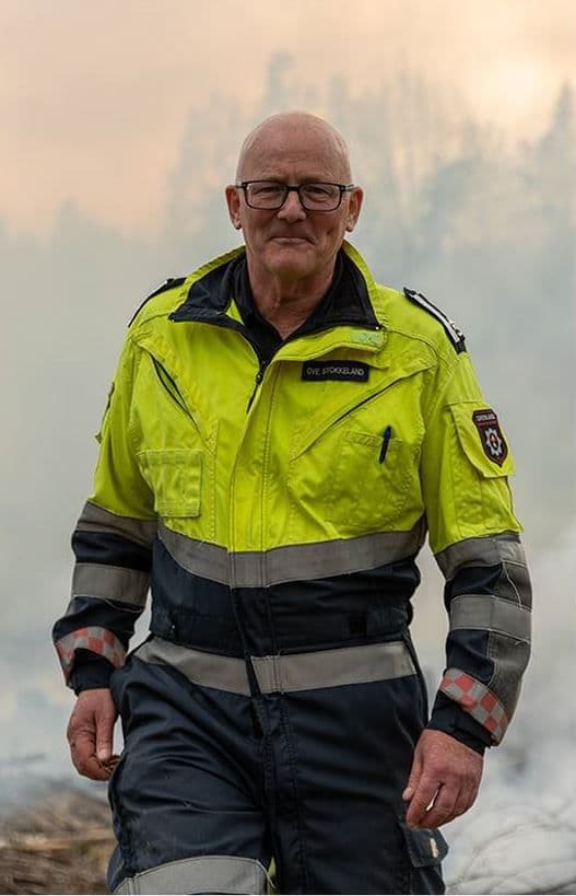 En skallet mann med briller, ved navn Ove Stokkeland, i en neongul og mørkeblå refleksuniform, står mot en disig, røykfylt bakgrunn.